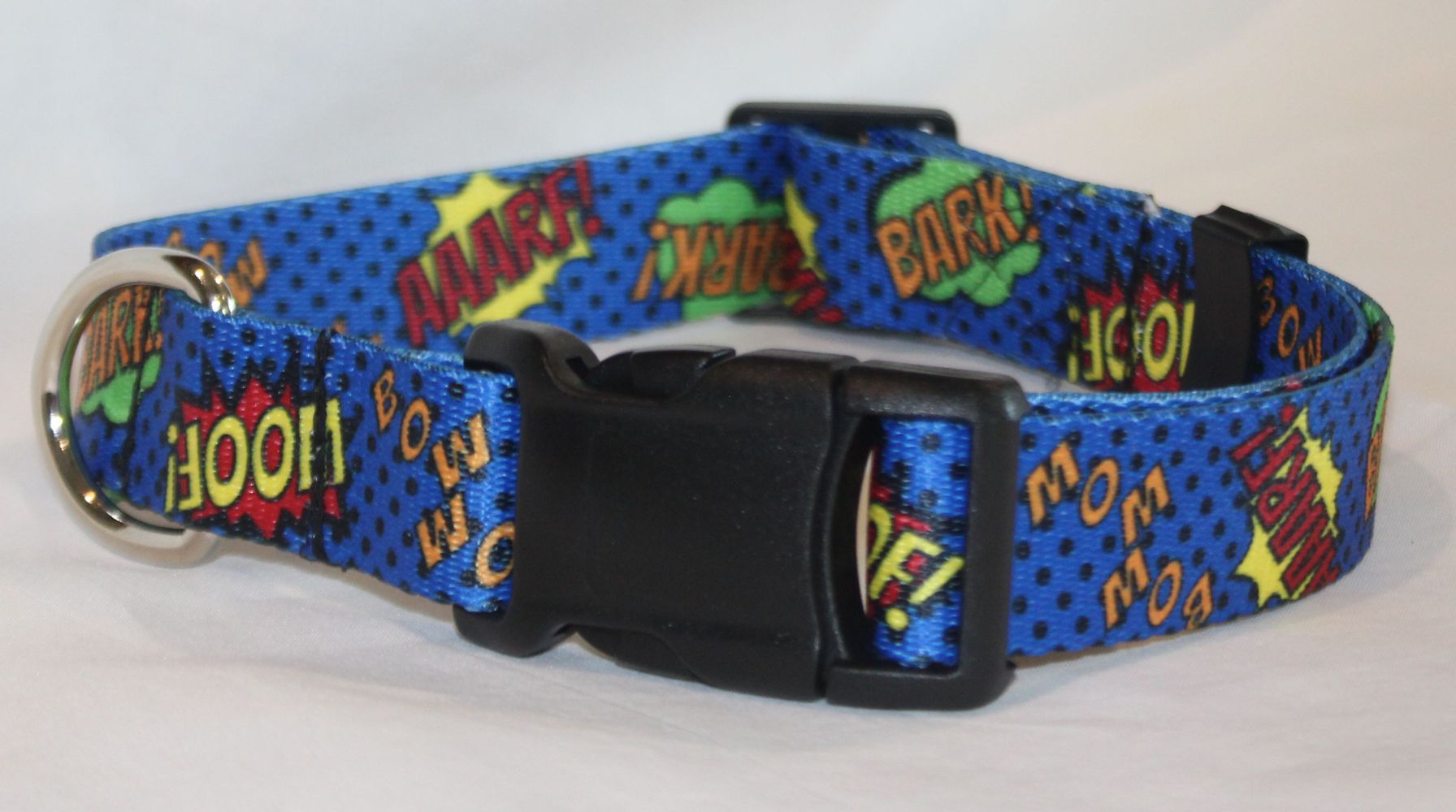 A blue dog collar with a comic book style print featuring pop-art text like 'WOOF!', 'BARK!', 'BOW!', and 'AARF!' in red, yellow, green, and white, with a black plastic buckle and metal ring.