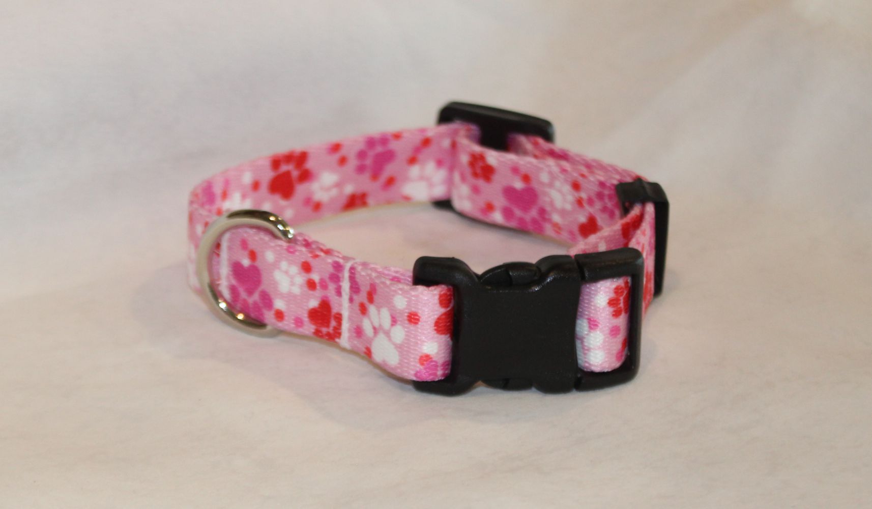 A pink nylon dog collar with a red and white paw pattern, featuring a black plastic quick-release buckle and a silver metal ring.