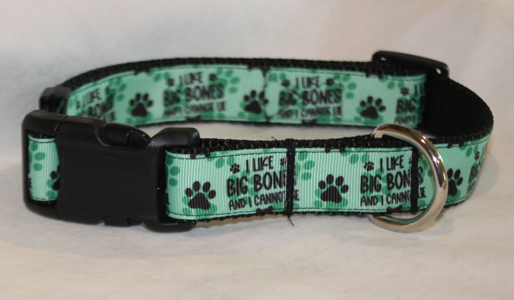 A pet collar with a mint green and black pattern featuring paw prints and the text 'I LIKE BIG BONES AND I CANNOPT LIE'. The collar has a black plastic buckle and a silver metal ring, 