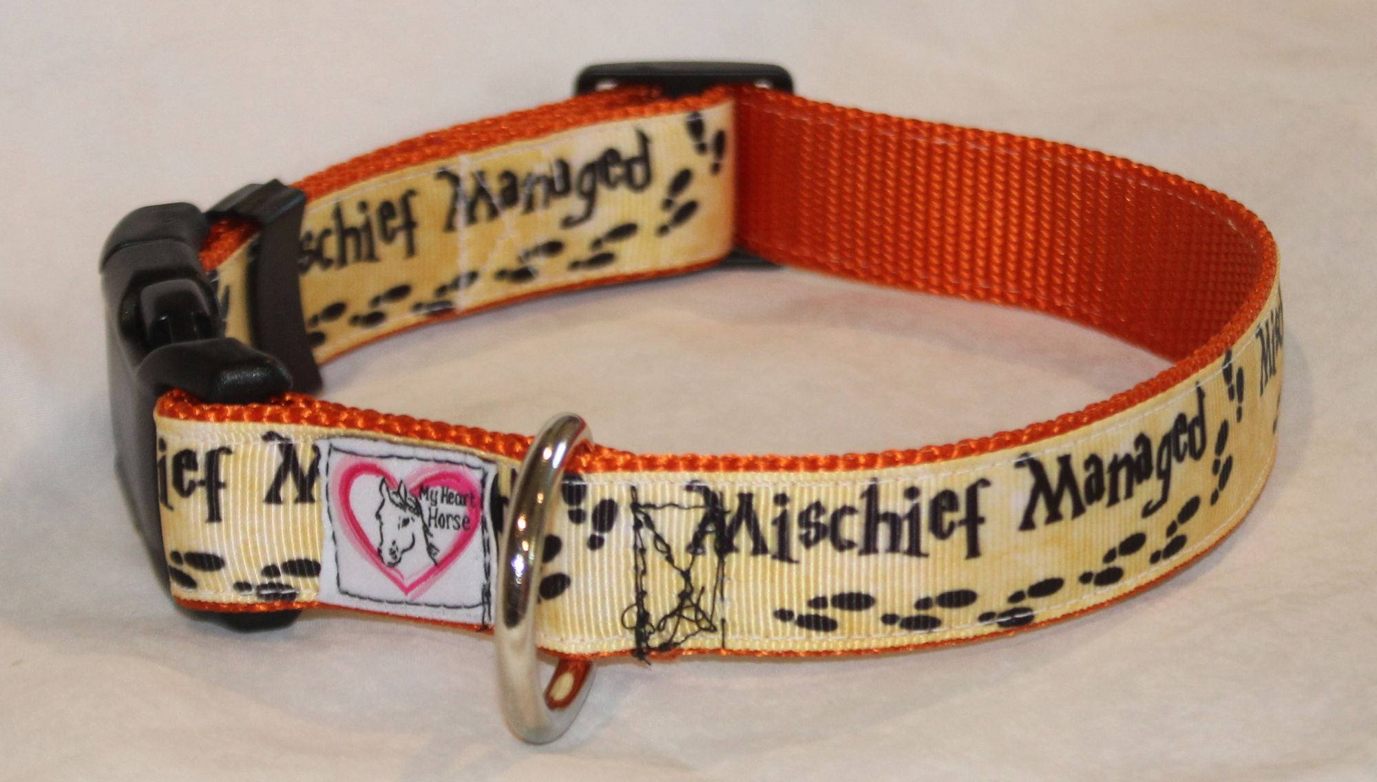 A dog collar with a cream-colored printed strap featuring mischief managed and paw prints, an orange inner band, and a black plastic buckle.