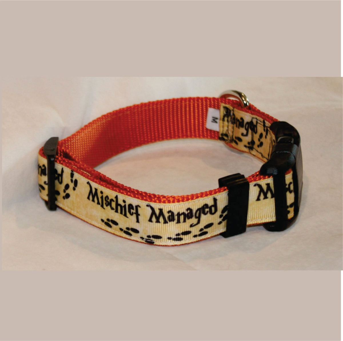 A dog collar with a cream-colored printed strap featuring the text 'Mischief Managed' in black gothic font and black paw prints, paired with a orange woven webbing band and black plastic hardware.