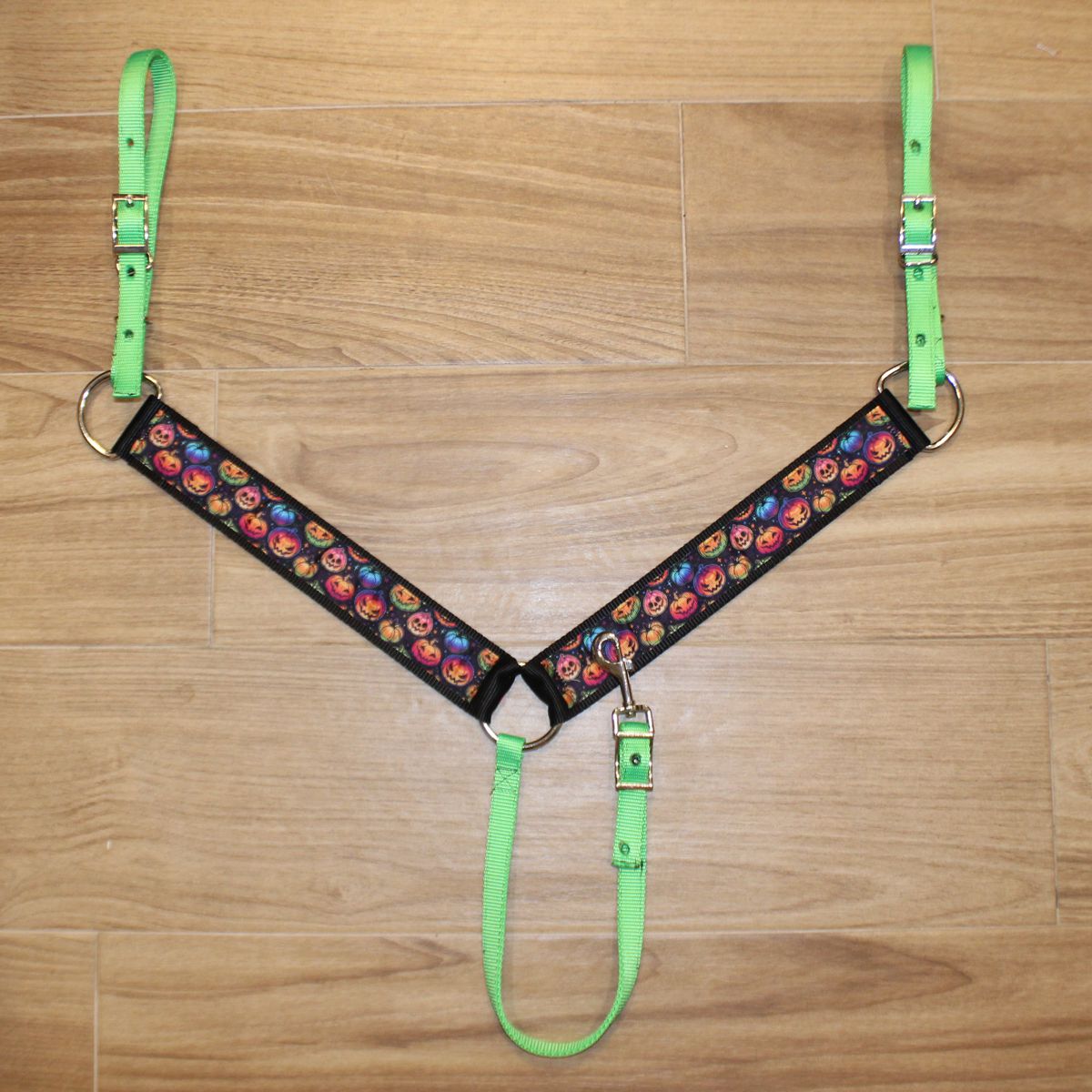 A horse breastcollar laid flat on a light brown wood floor, featuring bright neon green straps and a black central panel with a colorful pumpkin pattern.