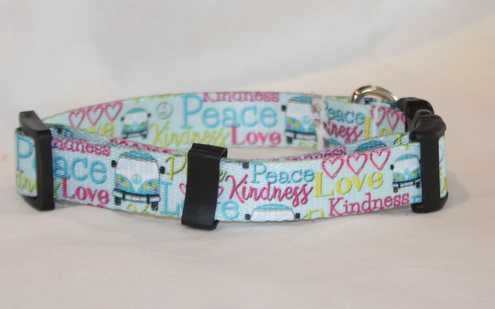 A nylon  dog collar with a colorful pattern of text reading 'Peace', 'Love', and 'Kindness' alongside small illustrations of vans and hearts, featuring black plastic buckles on a light background.