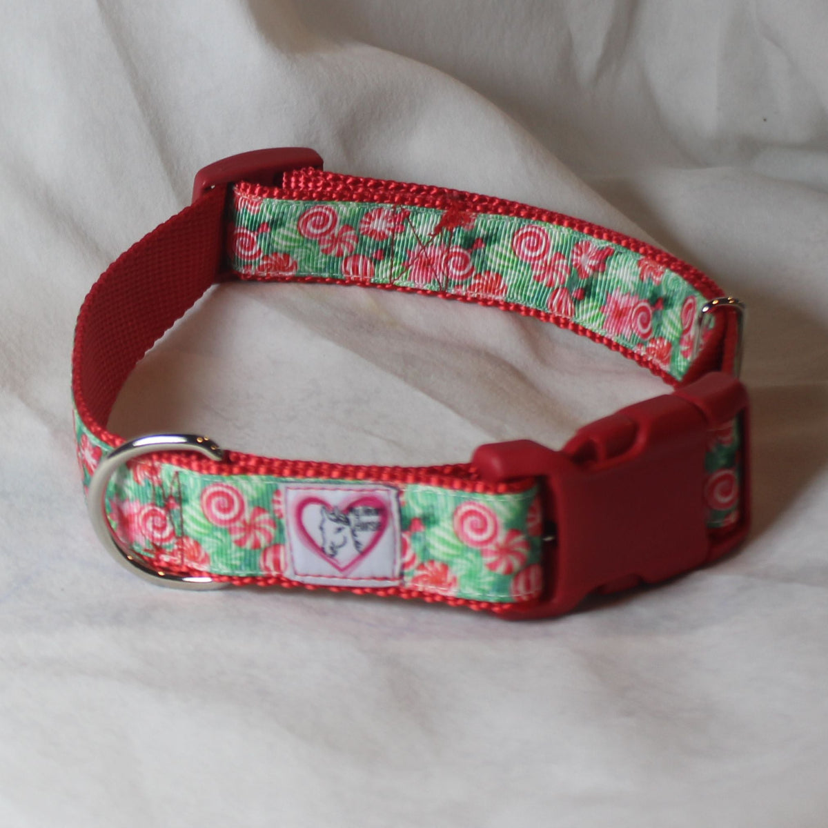 A red and green patterned dog collar with a red plastic buckle. The collar features a red and green candy cane pattern, a silver metal D-ring.l 