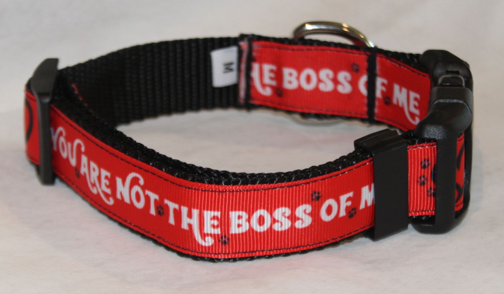 A red and black dog collar with the text 'YOU ARE NOT THE BOSS OF ME' printed in white, featuring black plastic buckles and a silver metal ring.
