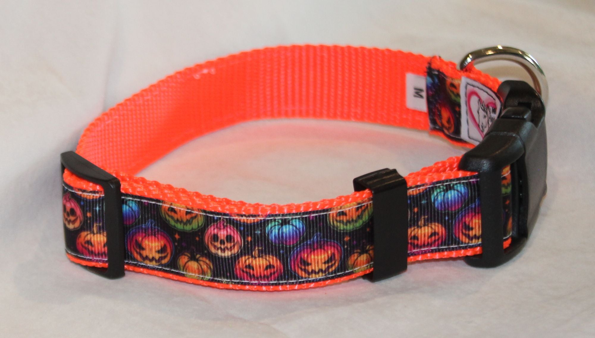 A bright orange dog collar with a black and multicolor pumpkin print strap, black plastic buckle, and a silver metal ring